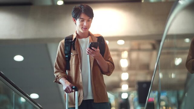 Young Asian man with backpack and wheeled suitcase checking smartphone while riding escalator inside modern airport terminal, male traveler using mobile phone during journey, air travel, tourism and d
