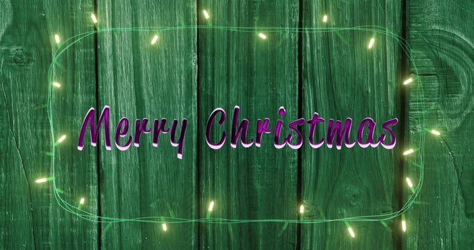 Glowing purple cursive Merry Christmas lettering sitting on green wooden planks, with fairy lights