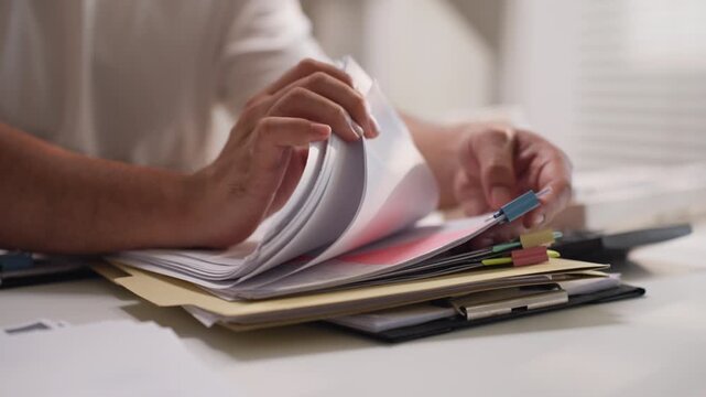 Asian office worker flipping through pages of paper document file rapidly at desk, dealing with heavy workload and deadline stress.