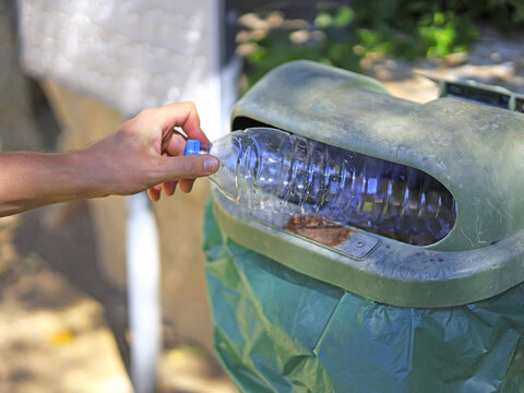 A hand pushes a plastic bottle through the opening of an outdoor bin, placing discarded drink packaging in public waste collection for municipal sanitation and proper disposal that prevents litter.