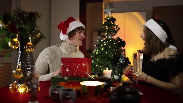 Christmas couple shares a playful moment as a smiling woman pulls down her partners Santa hat over his eyes while he laughs beside wrapped gifts and a glowing holiday tree