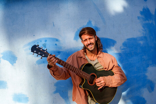Male musician smiling and strumming an acoustic guitar, bringing joyful energy and creative expression against a textured blue wall