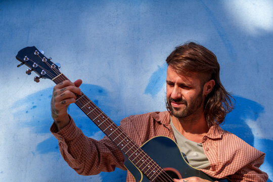Tattooed musician playing guitar in bright sunlight, with a blue textured wall framing a relaxed creative portrait