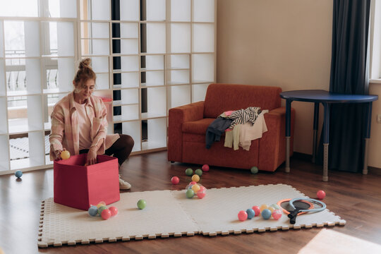 Woman seated beside pink storage box and scattered rainbow toys in sunlit apartment illustrates intentional downtime and balanced household routines after playful activity for mindful homemaking.