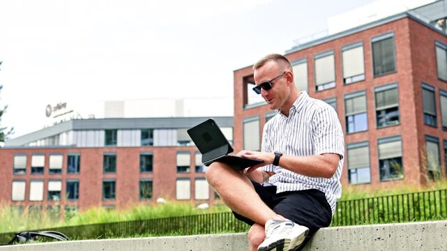 A man in sunglasses works on a laptop while sitting on a concrete ledge beside a green fence outside an office building during the day.
