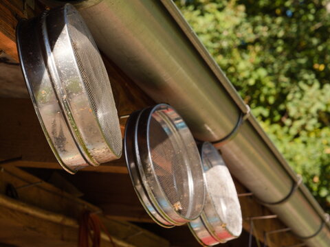 Metal honey strainers and sieves hanging under roof gutter on a bee farm, traditional beekeeping equipment for filtering raw honey, rustic apiculture tools outdoors on sunny day, close up view.