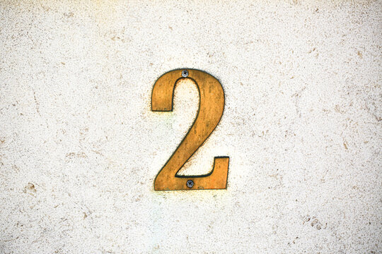 Number 2 vintage brass house number fixed with screws on rough plastered wall, aged golden metal digit with patina, address sign detail for real estate, architecture and street numbering concepts