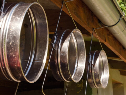 Professional stainless steel honey strainers hanging in a row on a bee farm, traditional beekeeping equipment for raw honey filtration, rustic apiculture tools, macro close up detail view.
