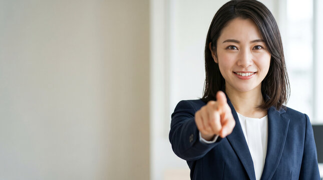 オフィスでカメラに向かって指差し爽やかに微笑むスーツ姿の日本人女性社員