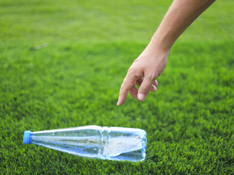 Hand reaching toward a plastic bottle on grass during litter cleanup, addressing waste management through responsible disposal of discarded drink packaging and reduction of outdoor plastic pollution.