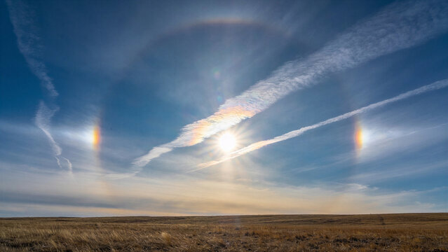 Radiant symmetrical sun dog rainbow phenomenon flanking the midday sun with contrails crossing above a dry golden steppe plain.