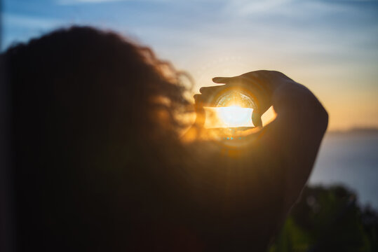 Back view silhouette of a woman holding a crystal ball or glass sphere to capture the golden sunset light over the sea. Vision, future perspective, and creativity concept. Human-created, not AI.