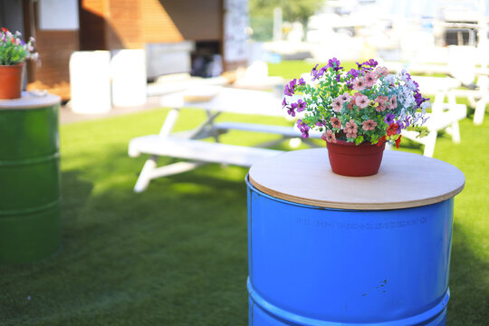 Colorful potted flowers top a blue barrel table in an outdoor seating area, pairing repurposed furniture with patio decor and a casual lounge layout framed by white sun loungers and artificial turf.