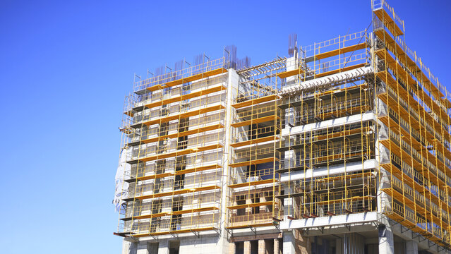 Unfinished concrete building wrapped in yellow scaffolding shows the structural stage of multistory construction, with exposed floors and reinforcing steel within a property development project.
