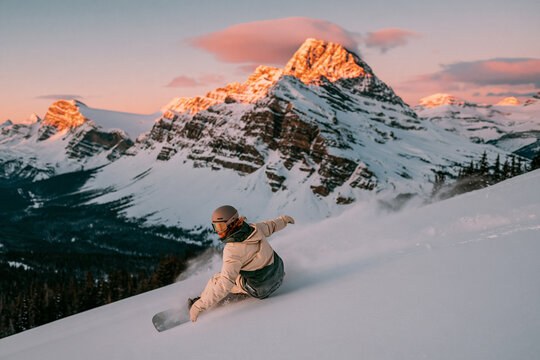 A snowboarder carves through deep powder on a steep mountain slope at golden hour with dramatic snow covered jagged peaks glowing in the soft sunset light in the background