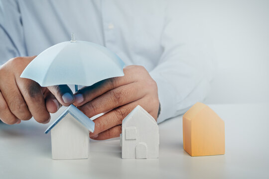 Businessman hands holding small toy umbrella over wooden house models on white desk, concept of home insurance, property protection, family safety, and risk management planning.