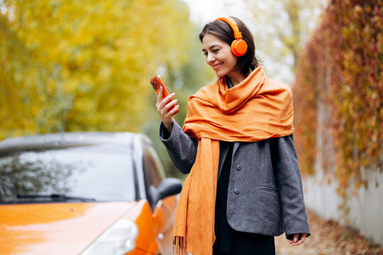 Woman wearing orange headphones and scarf, smiling while checking her smartphone beside a parked car on an autumn street