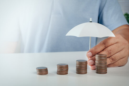 Hand holding small toy white umbrella over growing stack of coins on white table, concept of financial protection, money savings insurance, investment security, and risk management.