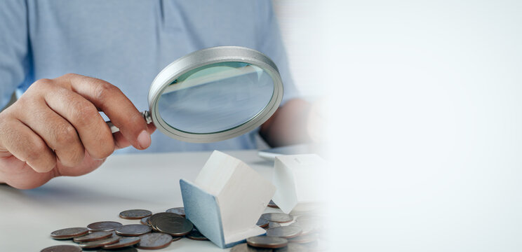 Businessman holding magnifying glass to inspect fallen wooden house model on stack of coins on white table, concept of house property inspection, real estate risk analysis, and mortgage crisis.