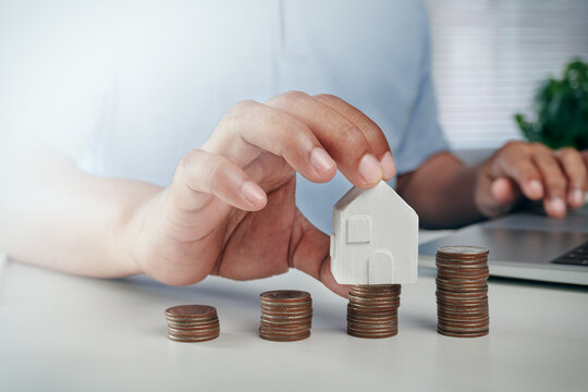 Businessman hand placing white house model on top of growing stack of coins on white desk, concept of house savings, real estate investment, property mortgage, and home loan planning.