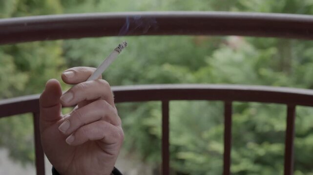A hand holds a cigarette beside a metal balcony railing, with green foliage beyond it during a steady outdoor shot