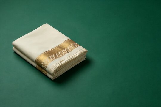 Traditional folded white Kerala mundu with gold border on a dark green background