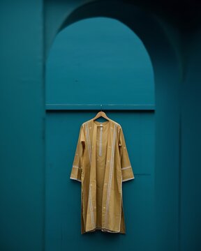 Traditional ochrecolored long embroidered cotton kurta hanging on a wooden hanger against a solid tealcolored arched wall