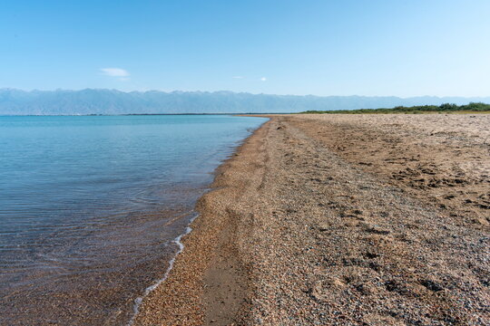 Empty wild pebble and sand beach along the clear shallow water of eastern Issyk Kul with distant mountains in the morning, Kyrgyzstan.