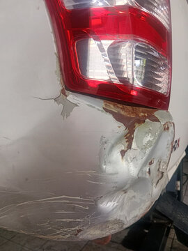 Close up view of a severely dented white car bumper with peeling paint and rust near the tail light