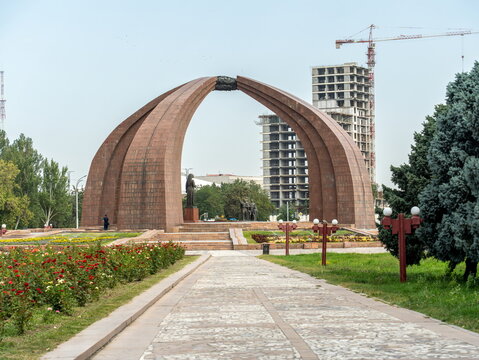 Red granite Victory Monument arch with rose beds and a paved path in Victory Square, Bishkek, Kyrgyzstan, with a high-rise under construction behind.
