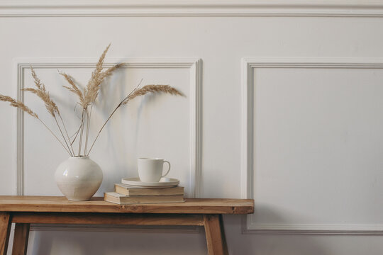 Minimalist Scandinavian Home Interior with White Ceramic Vase, Dried Festuca Grass, Coffee Cup and Vintage Books on Rustic Wooden Console Table. Neutral Nordic Decor, Elegant Wall Molding, Copy Space