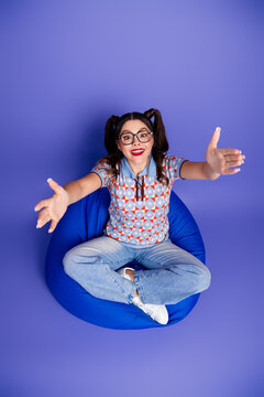 Young woman with glasses sits on blue beanbag reaching out with open arms smiling brightly