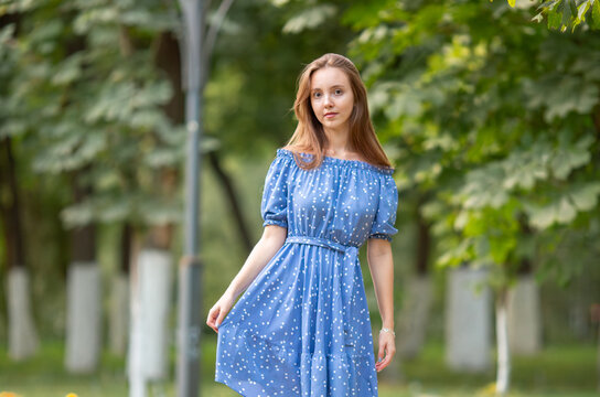A young smiling woman strolls through a spring park in a good mood. A beautiful young girl with long light brown hair in a dress poses among plants, trees, and flowers.