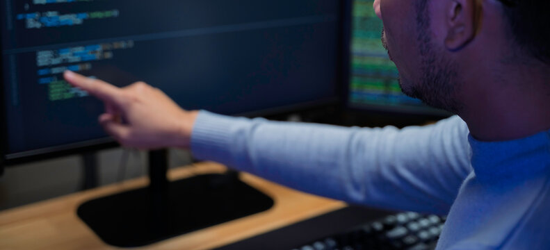 Computer programmer works at a multi monitor workstation in a dark office, reviewing programming code for software development, technical analysis, and system monitoring.