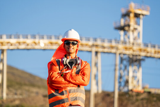 Mining worker in protective gear, standing with crossed arms before an industrial conveyor