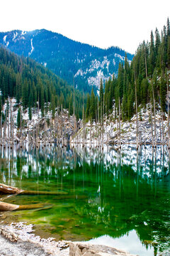 The flooded forest of Lake Kaindy in winter. A beautiful winter mountain landscape in Kazakhstan. A blue lake with tree trunks protruding from it against a backdrop of snow-capped mountains.