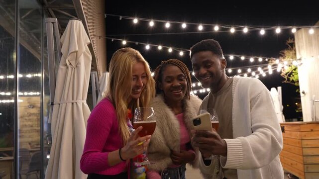 Cheerful multiracial friends holding drinks and taking selfies with a smartphone while celebrating at outdoor restaurant party