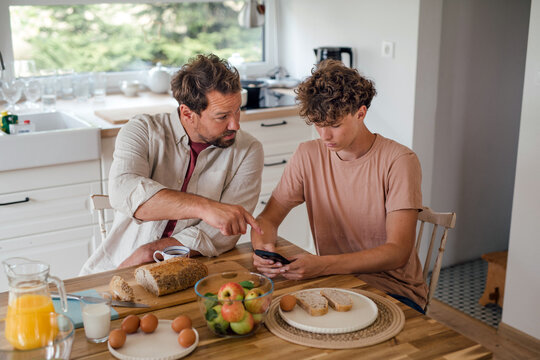 Father guiding teenage son using smartphone at breakfast table.