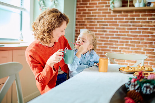 Mother and young daughter sharing a playful holiday breakfast, smiling together as the child enjoys a candy cane beside mugs and pastries