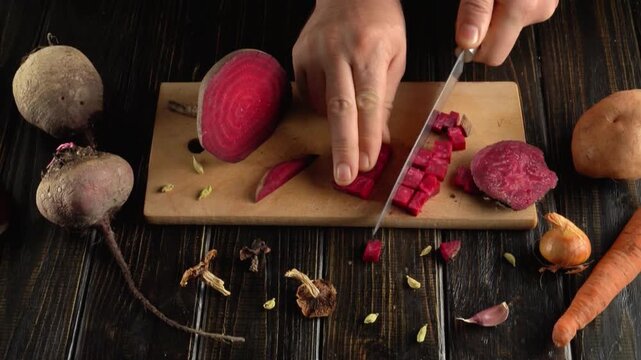 Hands slicing and dicing fresh red beet on wooden cutting board surrounded by root vegetables and scattered spices in a rustic kitchen preparation scene