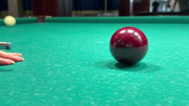 Wooden cue stick aims to strike dark red billiard ball on green felt cloth table inside sports club. Hand of player struck precise shot during Russian pyramid match, active motion.