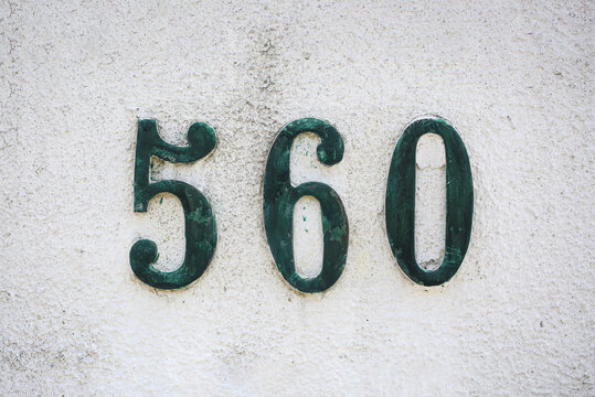 Number 560 in ornate vintage house numerals with verdigris patina mounted on cream plaster wall, antique address sign, aged metal digits, old building facade, textured stucco wall, European style