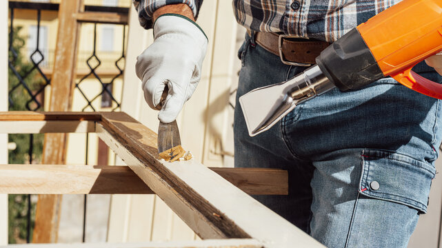 Adult carpenter wearing protective leather gloves, using a heat gun and a wood scraper to remove paint from an old wooden window. Construction industry, carpentry. Restoration. 