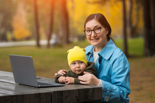 Smiling working mother sits outdoors holding baby boy beside open laptop computer on wooden picnic table. Cheerful female professional balances remote freelance parenting in autumn park.