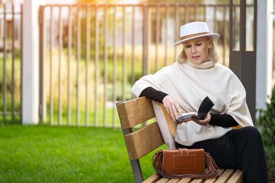 Older woman reading book on bench feels serene and stylish. Fashion lifestyle portrait shows woman in white hat, black gloves, and pale wrap, resting beside handbag in sunny outdoor space.