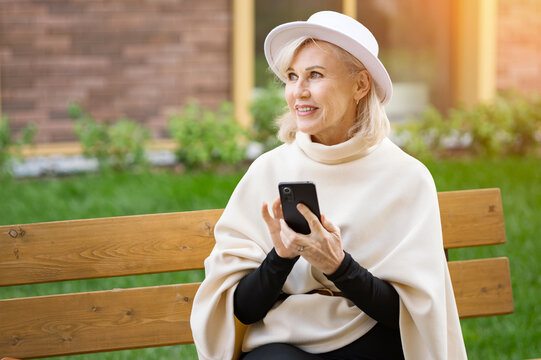 Happy older woman checks smartphone while sitting on wooden bench outdoors. Calm woman enjoys garden leisure in elegant white clothing, smiling beneath white hat in warm daylight, browsing mobile