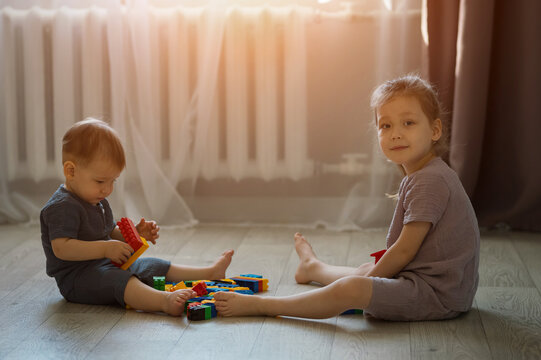 Young Siblings Assemble Plastic Building Blocks Across Home Living Room Floor. Sister Smiles Calmly Toward Camera While Focused Baby Brother Stacks Colorful Bricks During Quiet Daytime Play.