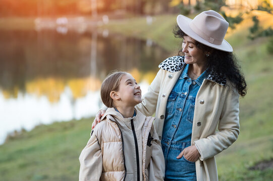 Smiling mother embraces happy daughter outdoors during peaceful family autumn lifestyle walk along lake shore, sharing cheerful moments together while dressed in stylish warm seasonal clothing.