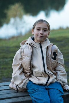 Smiling preteen child sits quietly on rustic wooden seat beside lake during autumn evening. Relaxed young girl keeps hands tucked in pockets while resting outdoors amidst serene natural park scenery.