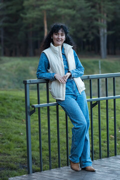 Content mature lady poses outdoors leaning against fence handrail on wooden walkway. Stylish person wears casual denim clothes and sleeveless padded jacket, looking relaxed in nature setting.